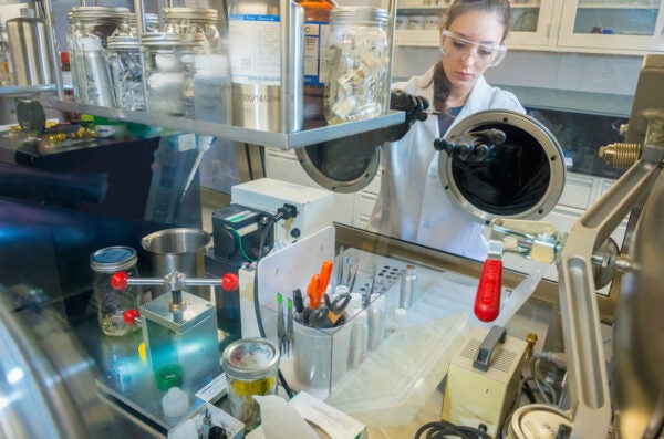 A female scientist in a lab coat pipettes behind lab equipment.