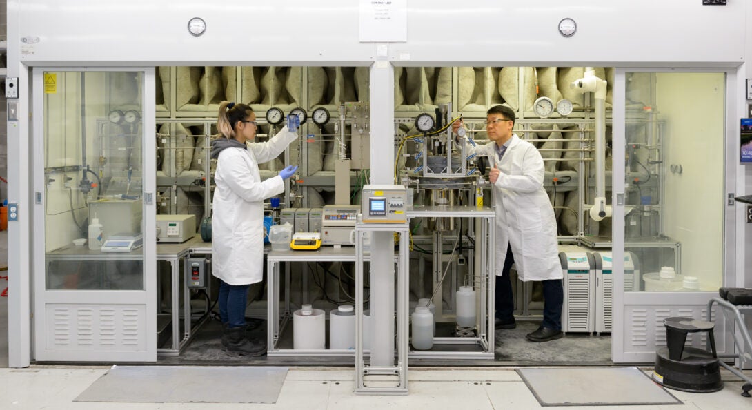 Two people in lab coats work on equipment separated by a vertical wall.