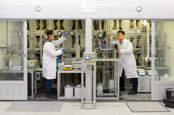 Two people in lab coats work on equipment separated by a vertical wall.