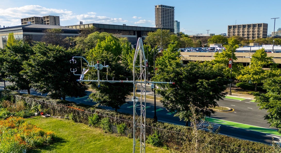 A metal tower with attached sensors in the foreground above a green field with UIC campus buildings in the background.